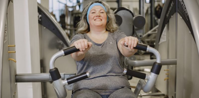 Smiling woman exercises on a gym machine, promoting fitness and well-being.